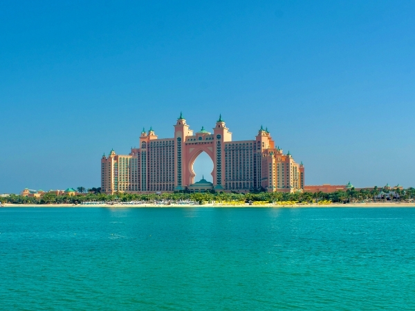 The Atlantis The Palm resort in Dubai stands prominently against a clear blue sky, framed by the tranquil waters of the Arabian Gulf with lush greenery at its base.