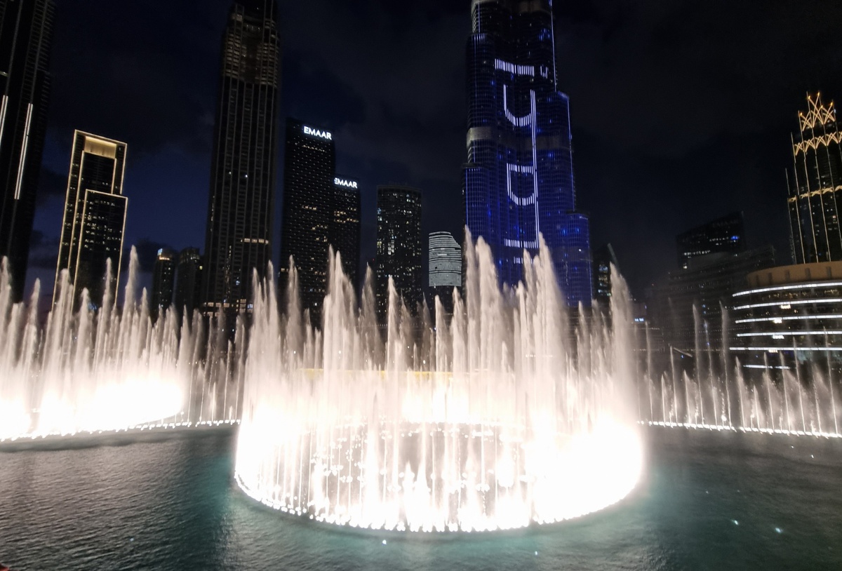 The Dubai fountains illuminated at night with the Burj Khalifa, Dubai's tallest tower (in a needle shape) in the background, with purple Arabic writing on it. 