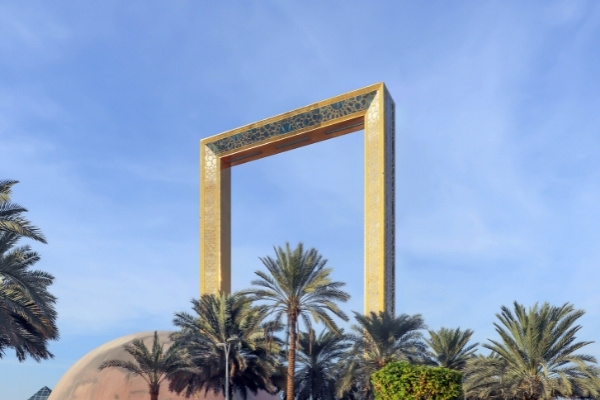 One of Dubai landmarks, the Frame, which looks like a giant gold picture frame. Below the frame are green palm trees. 