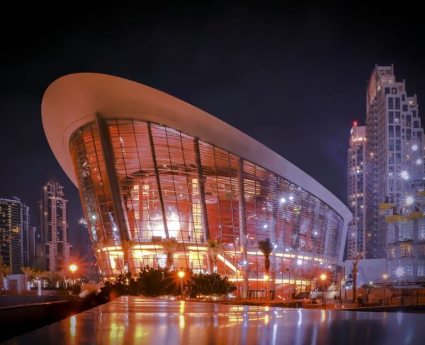 The sleek and modern Dubai Opera House illuminated at night, with its iconic dhow-shaped architecture, stands out against the backdrop of the city's glittering skyscrapers.