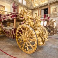 An ornate golden carriage with intricate sculptural details and red velvet accents on display in a museum, showcasing the grandeur of historical transportation."