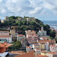 Scenic view of the historic São Jorge Castle perched atop a hill overlooking the colorful buildings of Lisbon, with the Tagus River and distant cityscape under a partly cloudy sky.