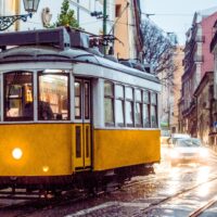 A yellow and white vintage tram makes its way down a rainy, cobblestone street in Lisbon, illuminated by its headlights and a soft glow from streetlights, with a car following behind and a serene cityscape in the background.